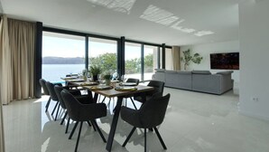 Uma sala de jantar moderna com uma mesa posta para quatro pessoas, grandes janelas com vista para o mar e um sofá cinza ao fundo.