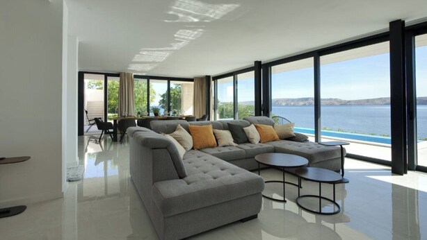 Uma sala de estar moderna com sofá em formato de L, duas mesinhas laterais e vista para o mar.