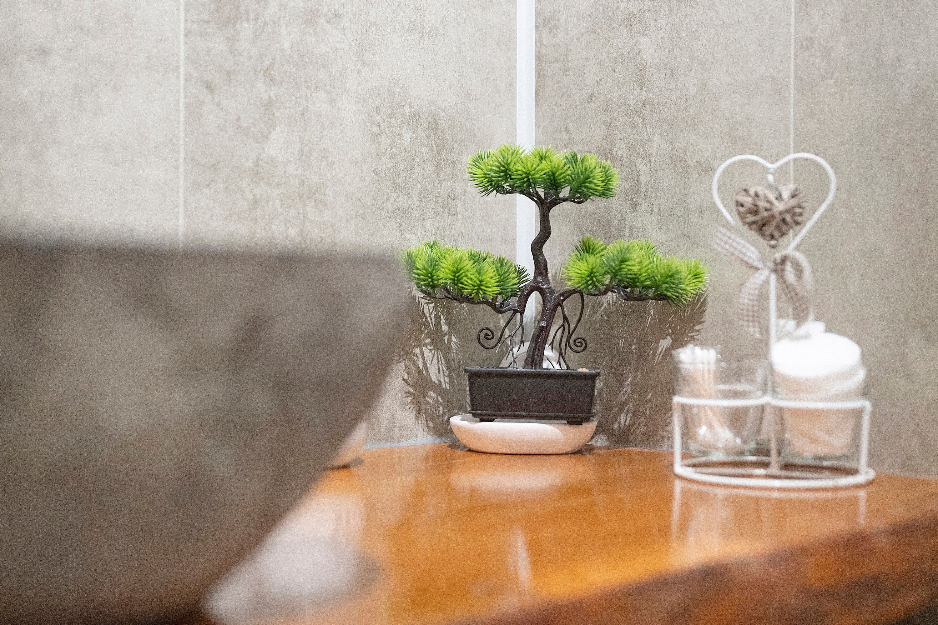 A bonsai tree in a black pot on a wooden table with a glass candle holder and a heart-shaped decoration.