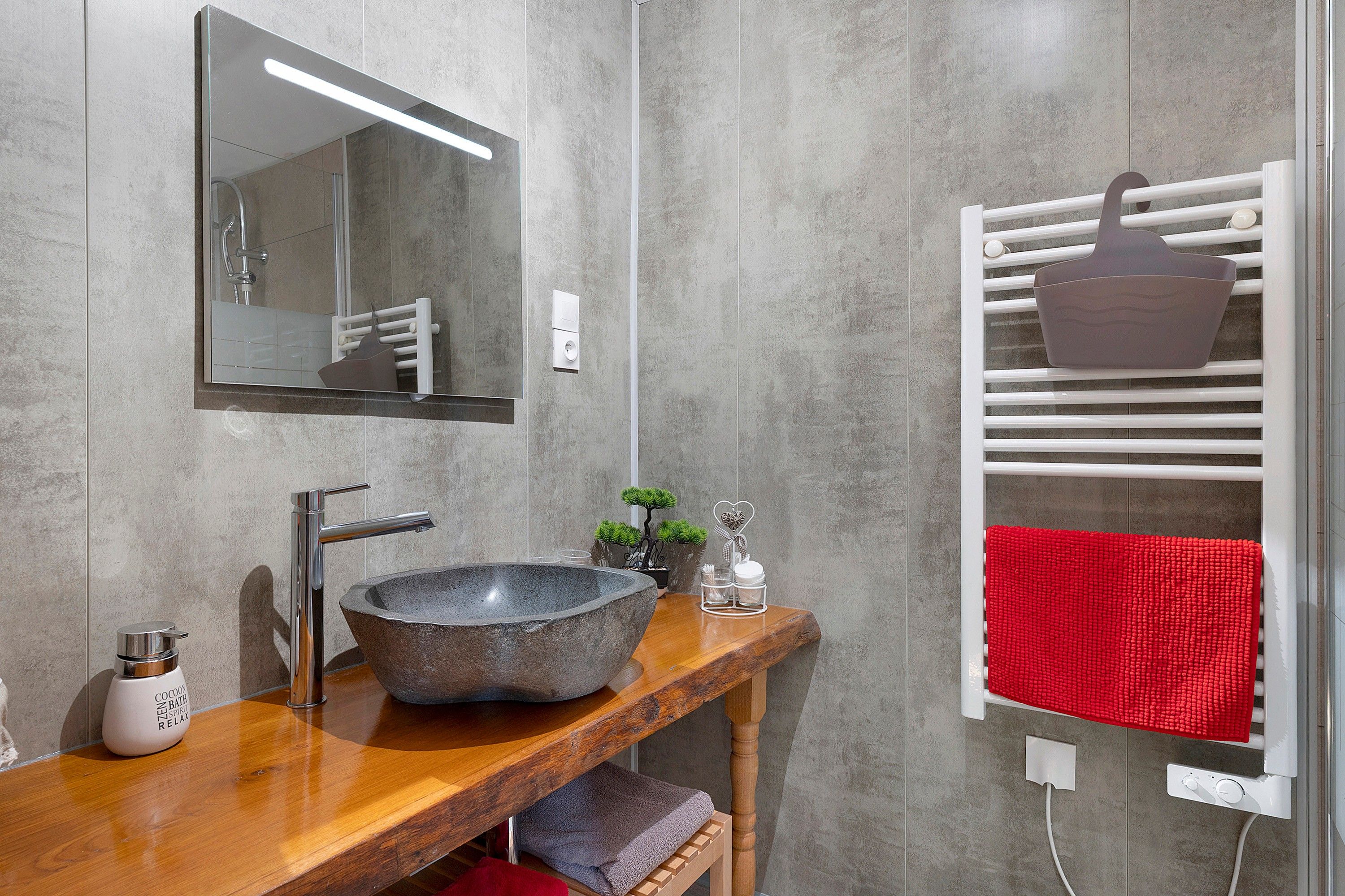 A modern bathroom with a stone sink, a wooden vanity, a mirror, a towel rack with a red towel, and a small plant.