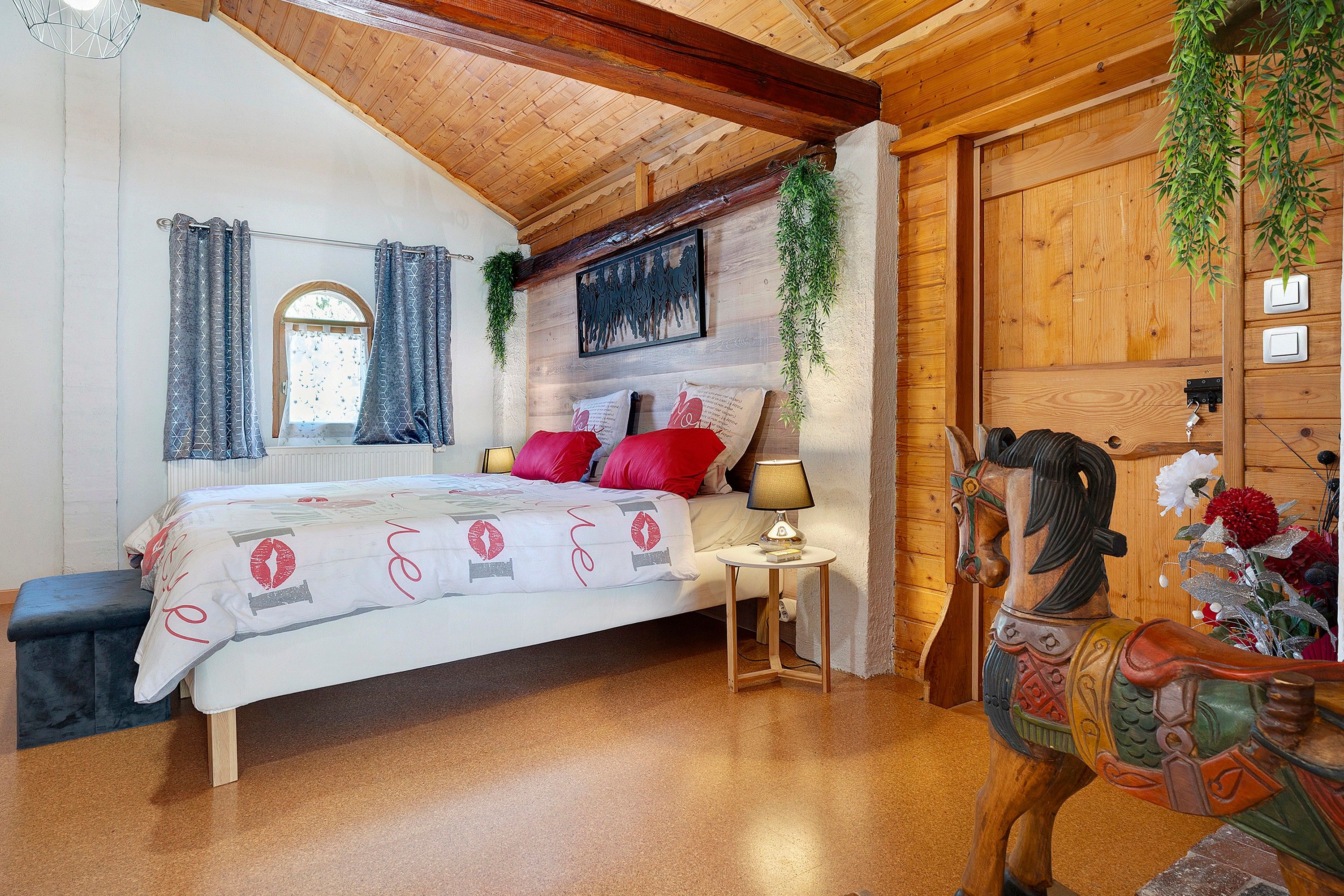 Een gezellig slaapkamer met een houten plafond, een bed met rode kussens, een klein nachtkastje met een lamp en een decoratief paardbeeldje.