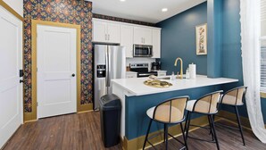A kitchen with white cabinets, a stainless steel refrigerator, a microwave, and a sink. There are three bar stools and a trash can. - The Bavarian Inn 5 Artsy Gem (Covington)