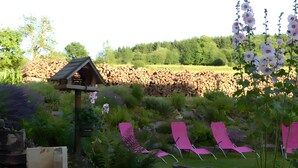 Een tuin met een vogelhuisje, een stapel houtblokken en roze stoelen.