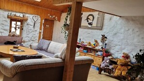 A cozy living room with a wooden ceiling, a fireplace, a sofa, a coffee table, and a bookshelf filled with stuffed animals. - Guesthouse “Goddess of the Harvest Room” with Shared Terrace, Garden, and Wi-Fi (Gerbépal)