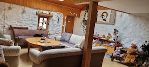 A cozy living room with a wooden ceiling, a fireplace, a sofa, a coffee table, and a bookshelf filled with stuffed animals.