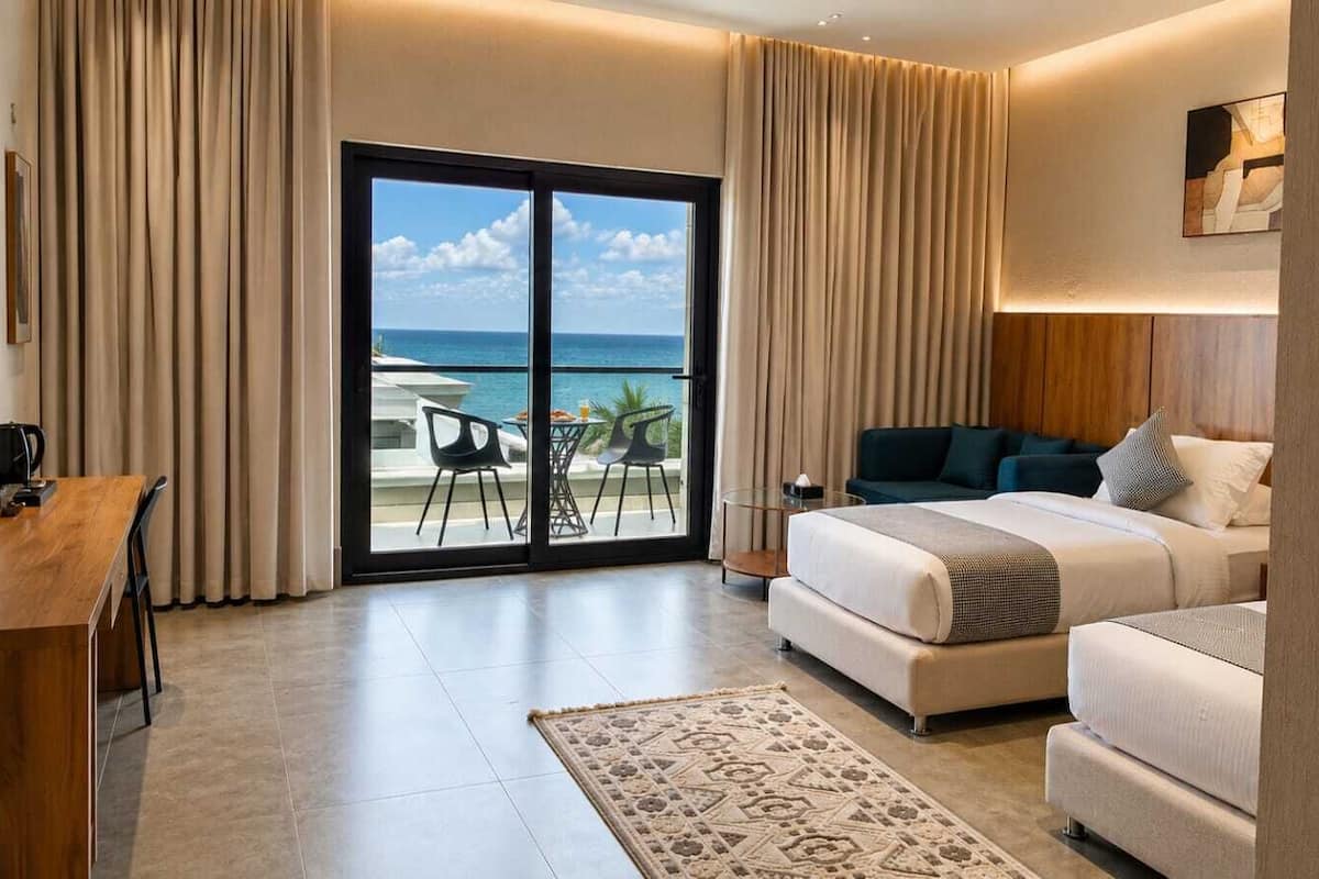 A hotel room with a bed, a desk, a chair, a view of the ocean, and a balcony.