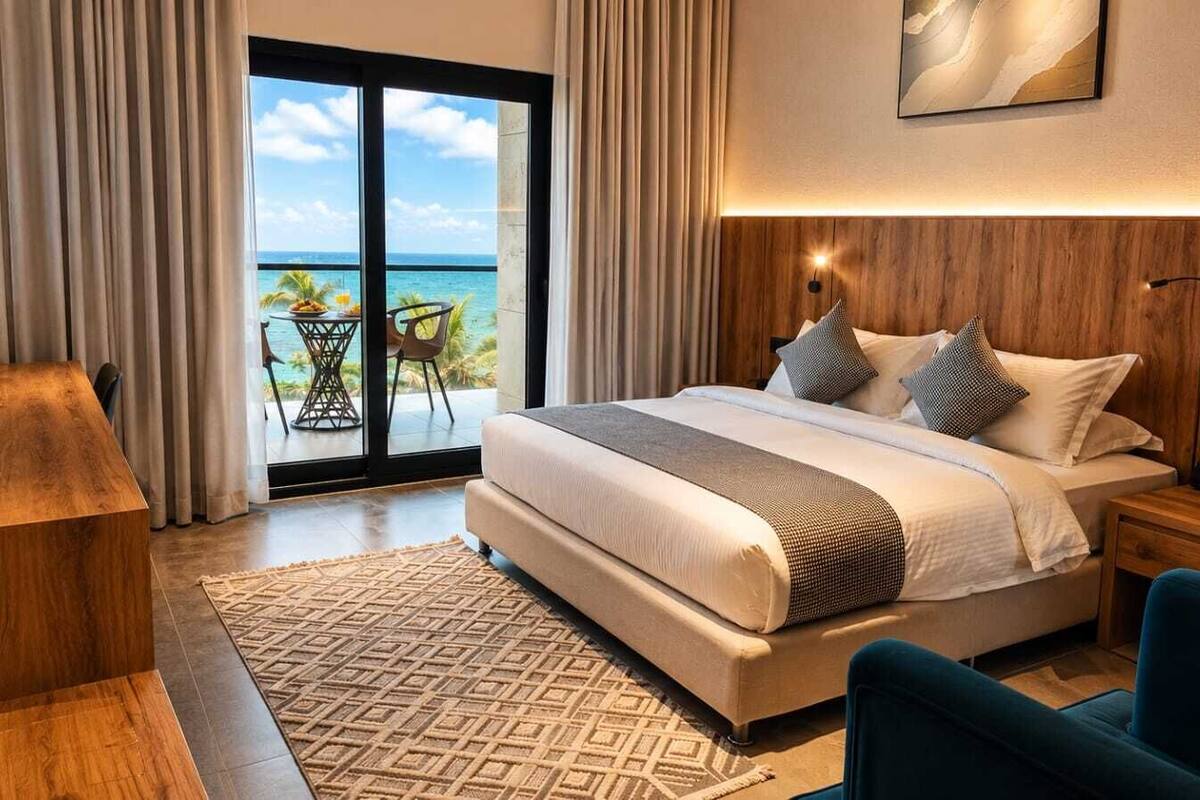 A bedroom with a large bed, a desk, a chair, a nightstand, and a view of the ocean.