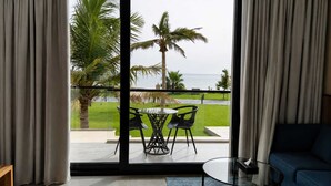 Una habitación con un gran ventanal que da a un balcón con mesa y sillas, un sofá azul y vistas al mar y palmeras.
