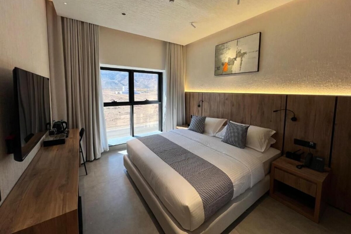 A modern hotel room with a large bed, a flat-screen TV, a desk, and a window with curtains.
