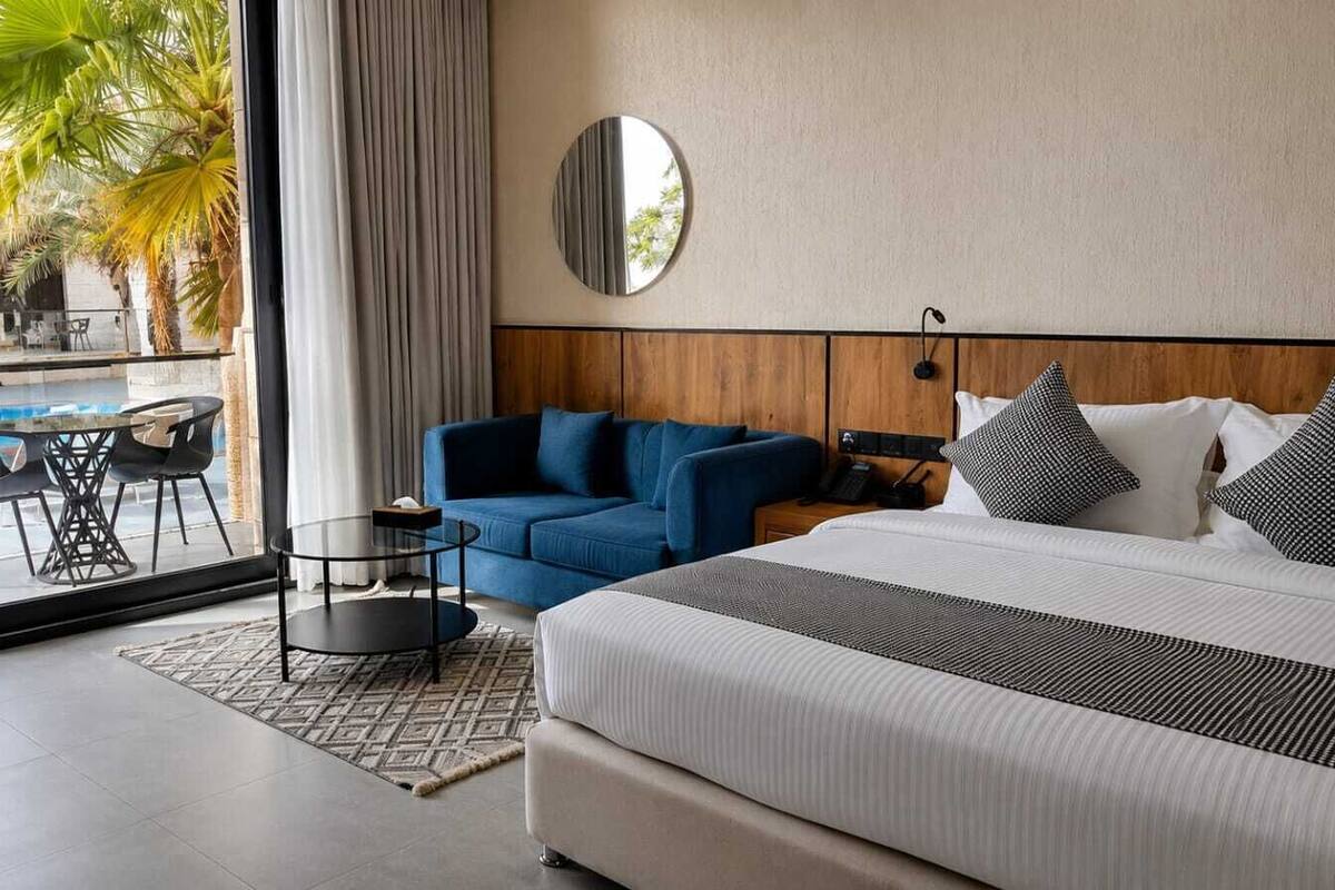 A modern hotel room with a large bed, a blue sofa, a round mirror, and a view of a pool and palm trees outside.