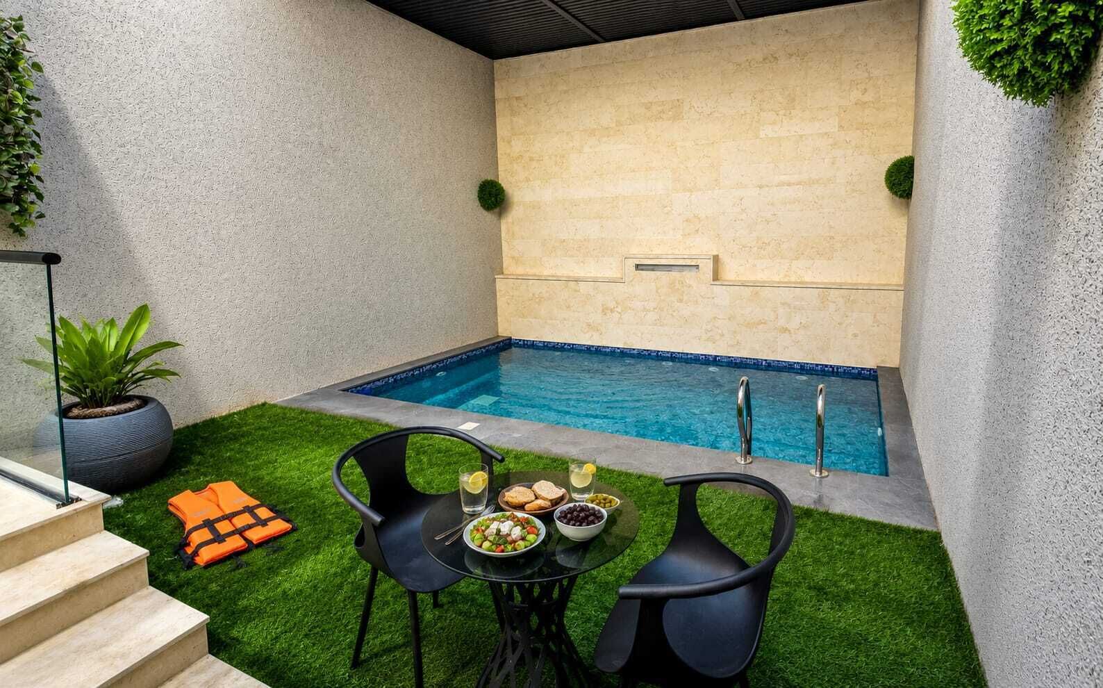 Zona de piscina con césped artificial, dos sillas, una mesa con comida y una escalera.