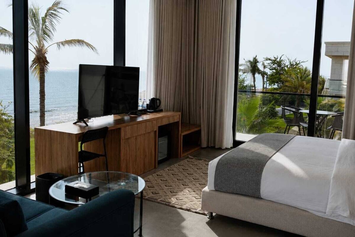 A hotel room with a bed, a desk, a TV, a balcony with a view of the sea, and a dining area.