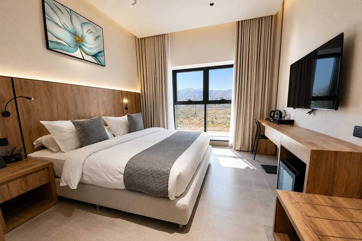 A modern hotel room with a large bed, a desk, a TV, and a view of the mountains.