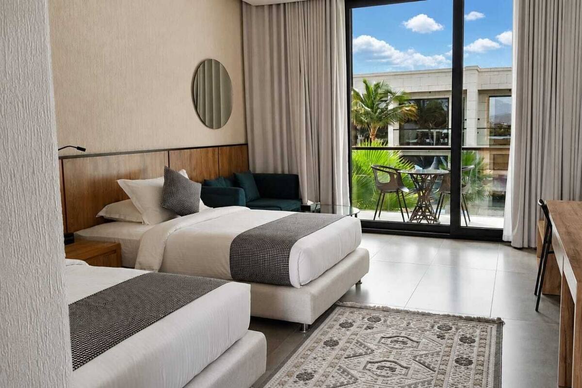 A hotel room with two beds, a balcony with a table and chairs, and a view of the outside.