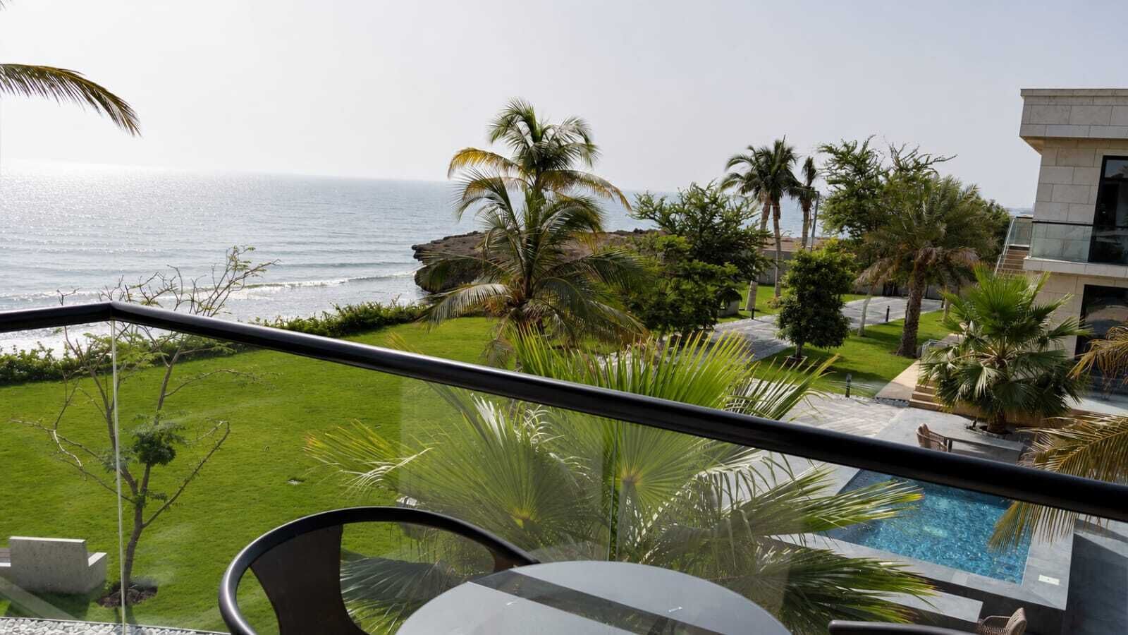 A balcony with a view of the ocean, a swimming pool, and palm trees.