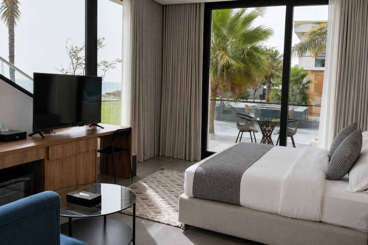 A hotel room with a bed, a desk with a TV, a chair, a small table, and a balcony with a view of palm trees and buildings.