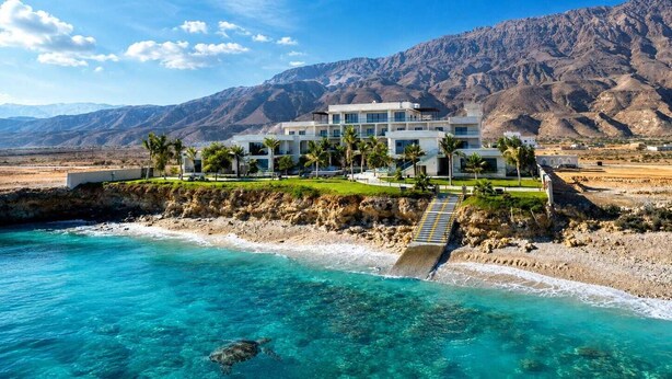 Un hotel frente al mar con diseño moderno, vistas a la playa y montañas al fondo.