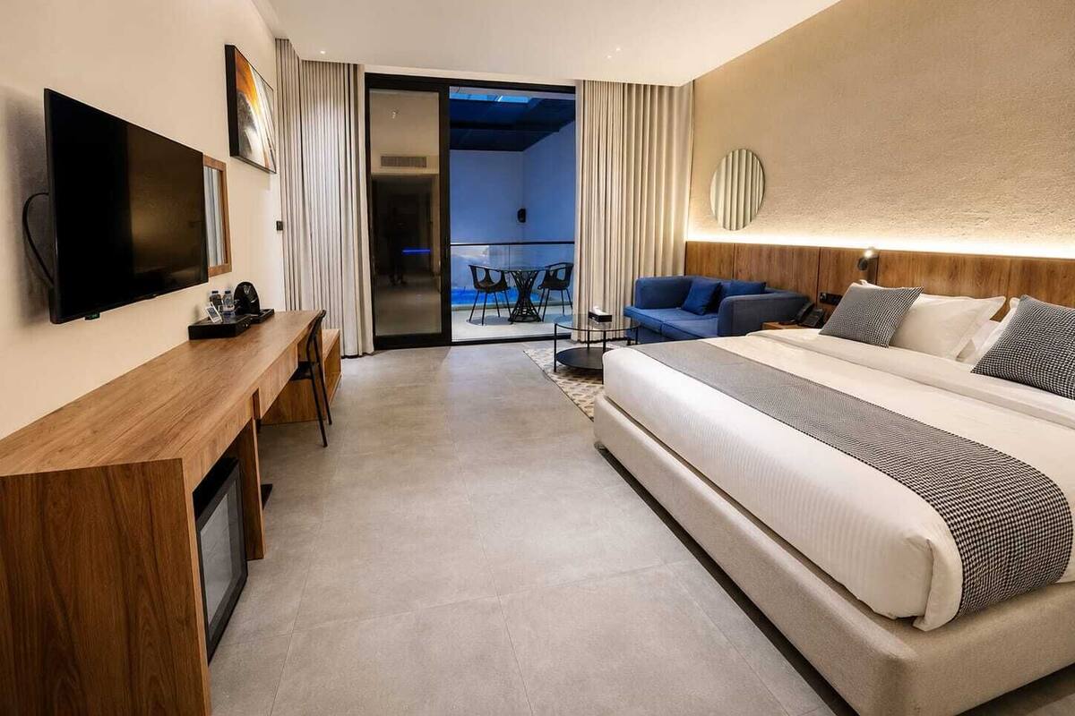 A modern hotel room with a large bed, a desk with a flat-screen TV, a balcony with a view, and a well-lit ceiling.
