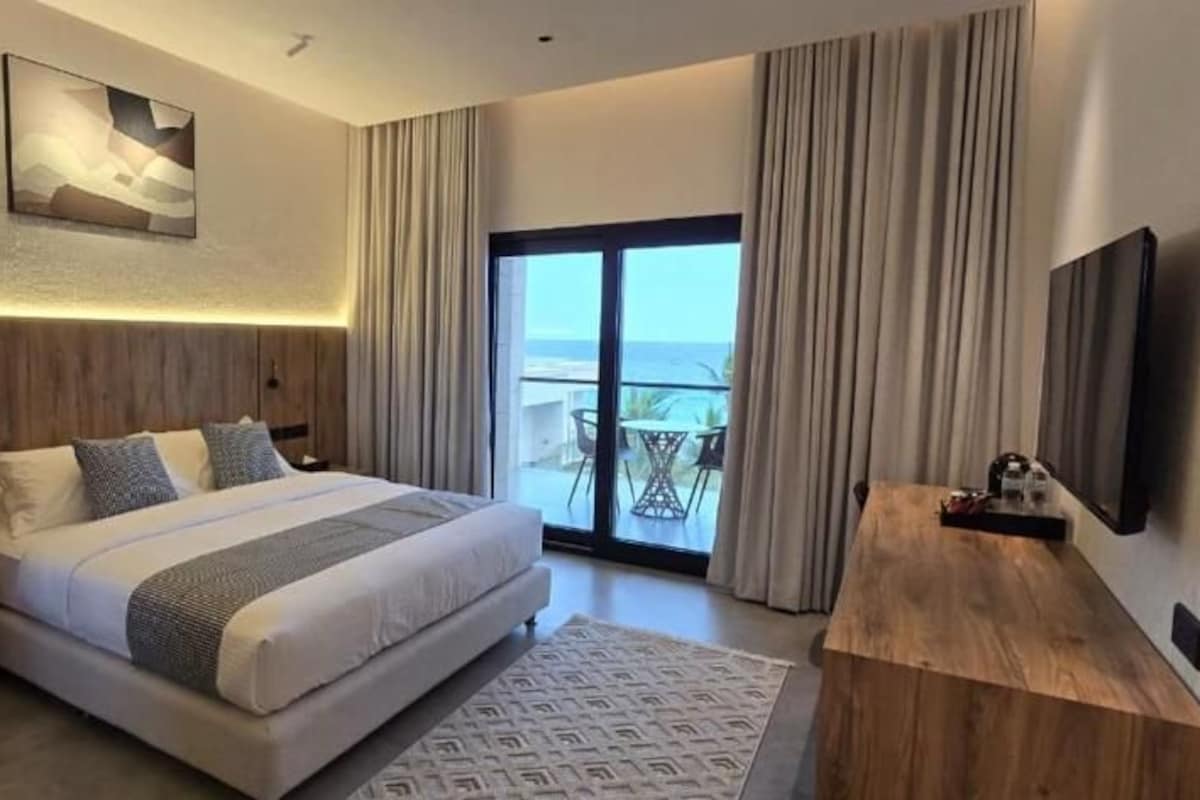 A modern hotel room with a large bed, a desk, a flat-screen TV, and a balcony with a view.