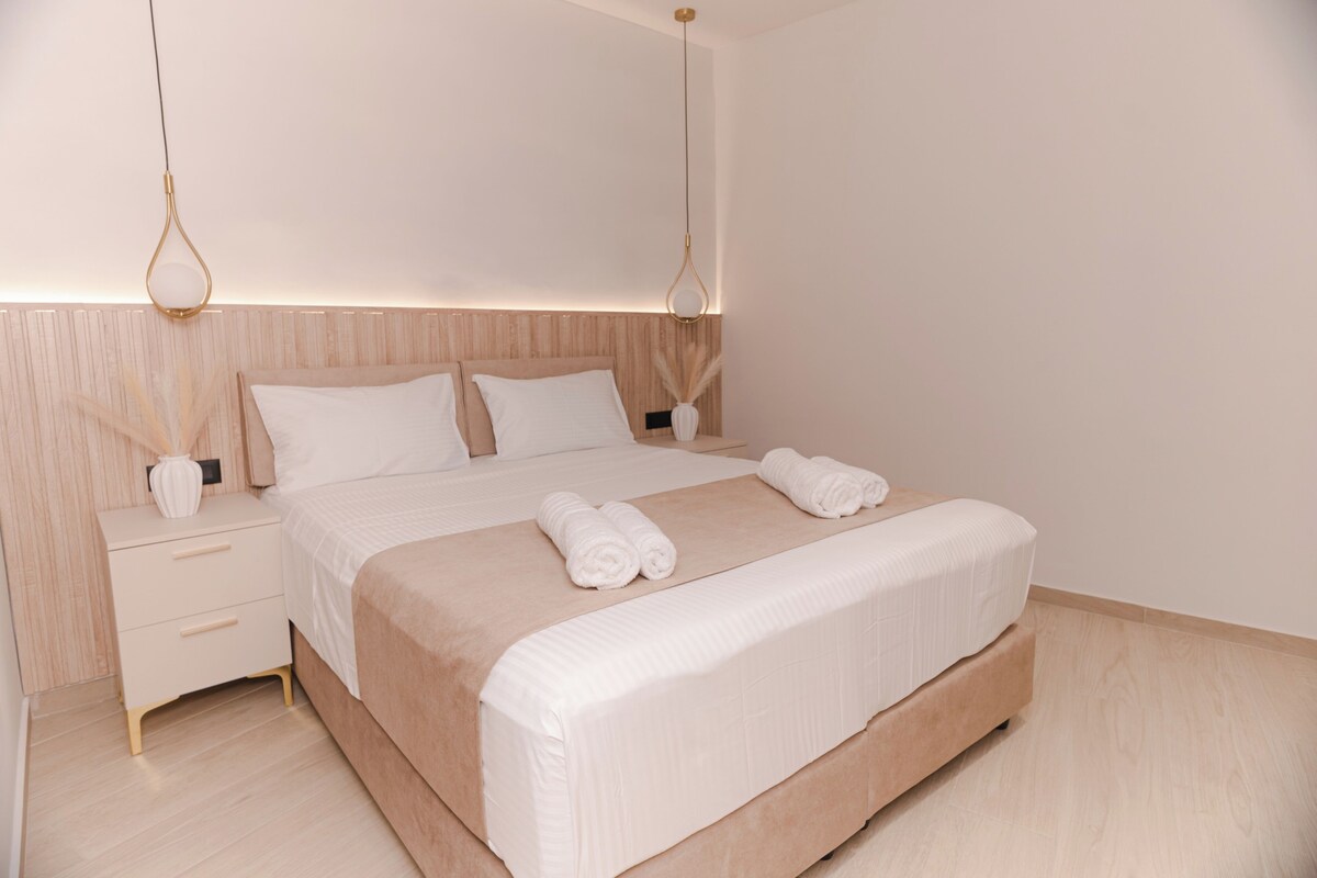 Una cama bien tendida con sábanas blancas y toallas enrolladas, una mesita de noche con un florero y una lámpara colgante.