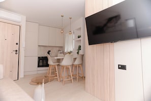 A modern kitchen with white cabinets, a dining area with a glass table and wooden chairs, and a mounted flat-screen TV.
