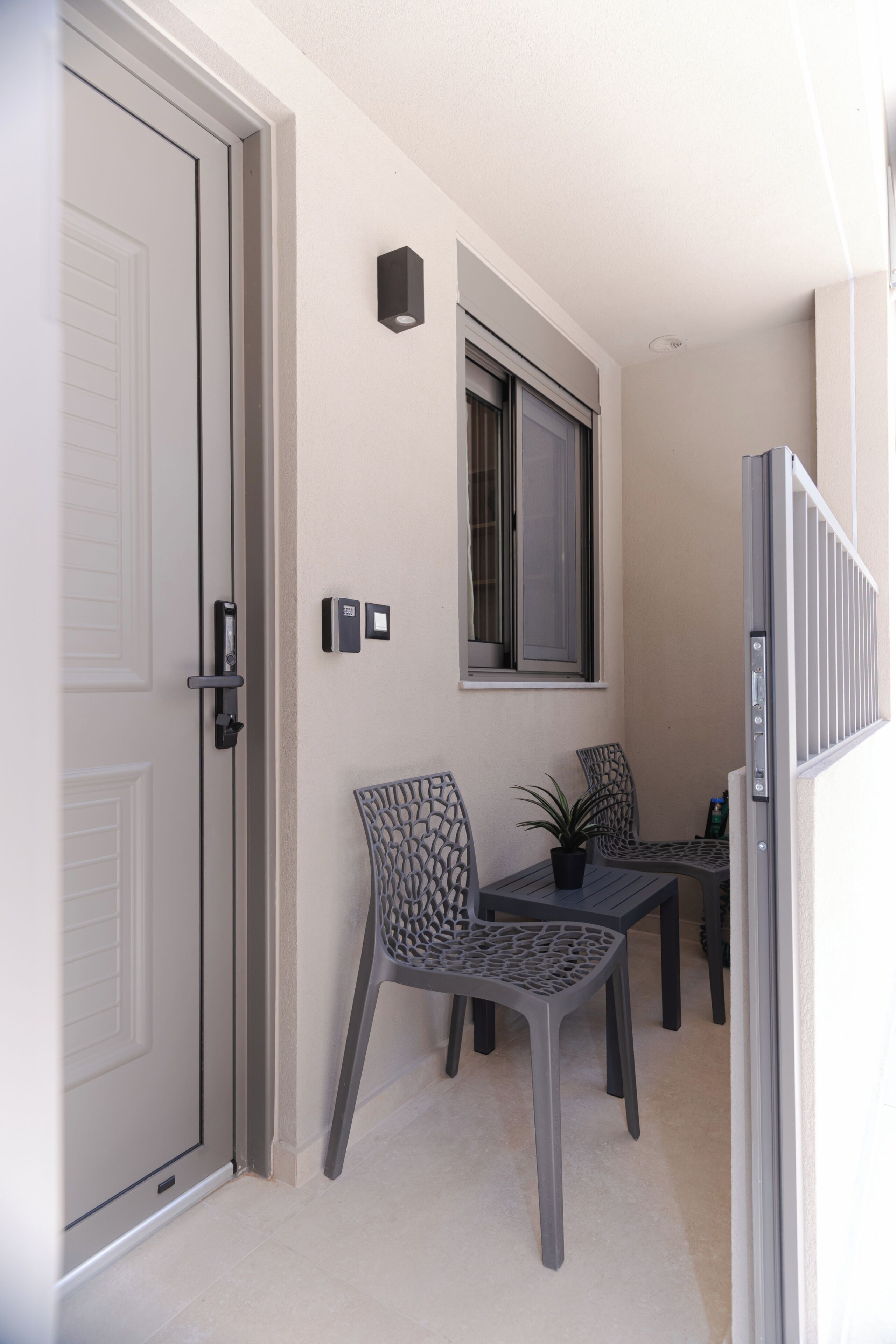 A room with a door, a window, a wall-mounted light, and two chairs with a potted plant.