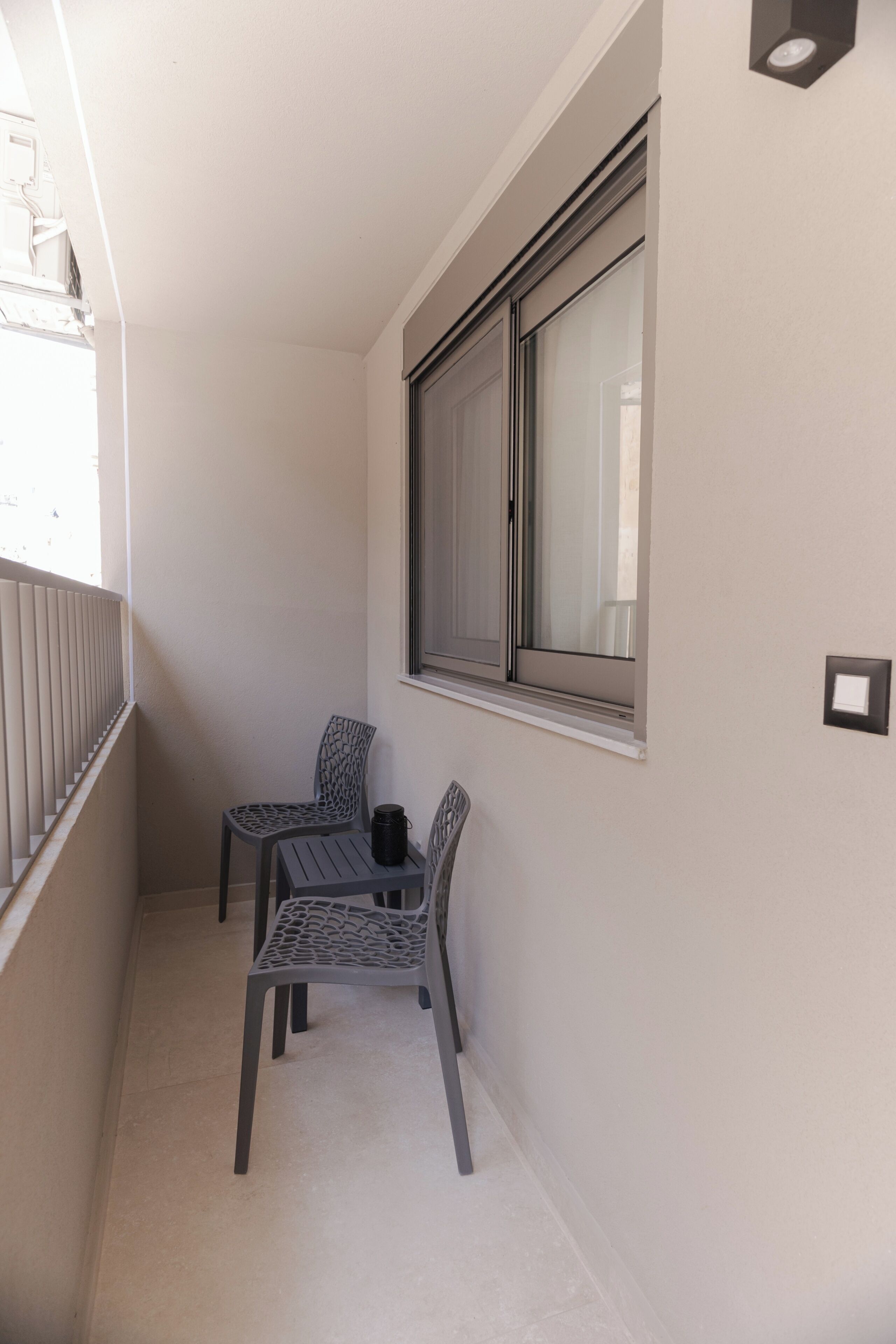 Ein schmaler Balkon mit zwei Stühlen und einem kleinen Tisch, ein Fenster mit Jalousien und einem Lichtschalter an der Wand.