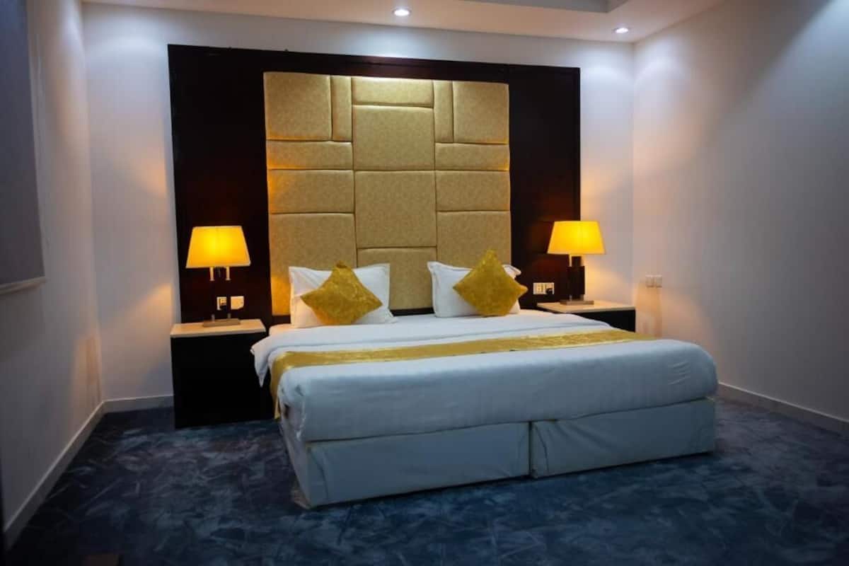Quarto de hotel com uma cama grande, duas luminárias de cabeceira, uma cabeceira e um tapete escuro.