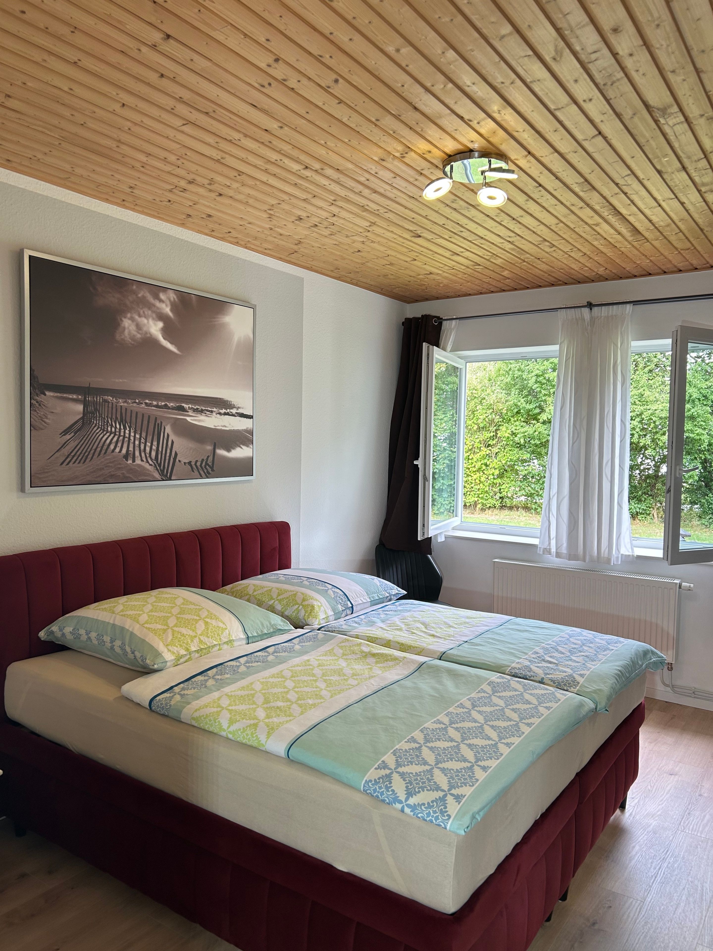 A bedroom with a wooden ceiling, a bed with patterned bedding, a large window with curtains, and a wall-mounted picture.