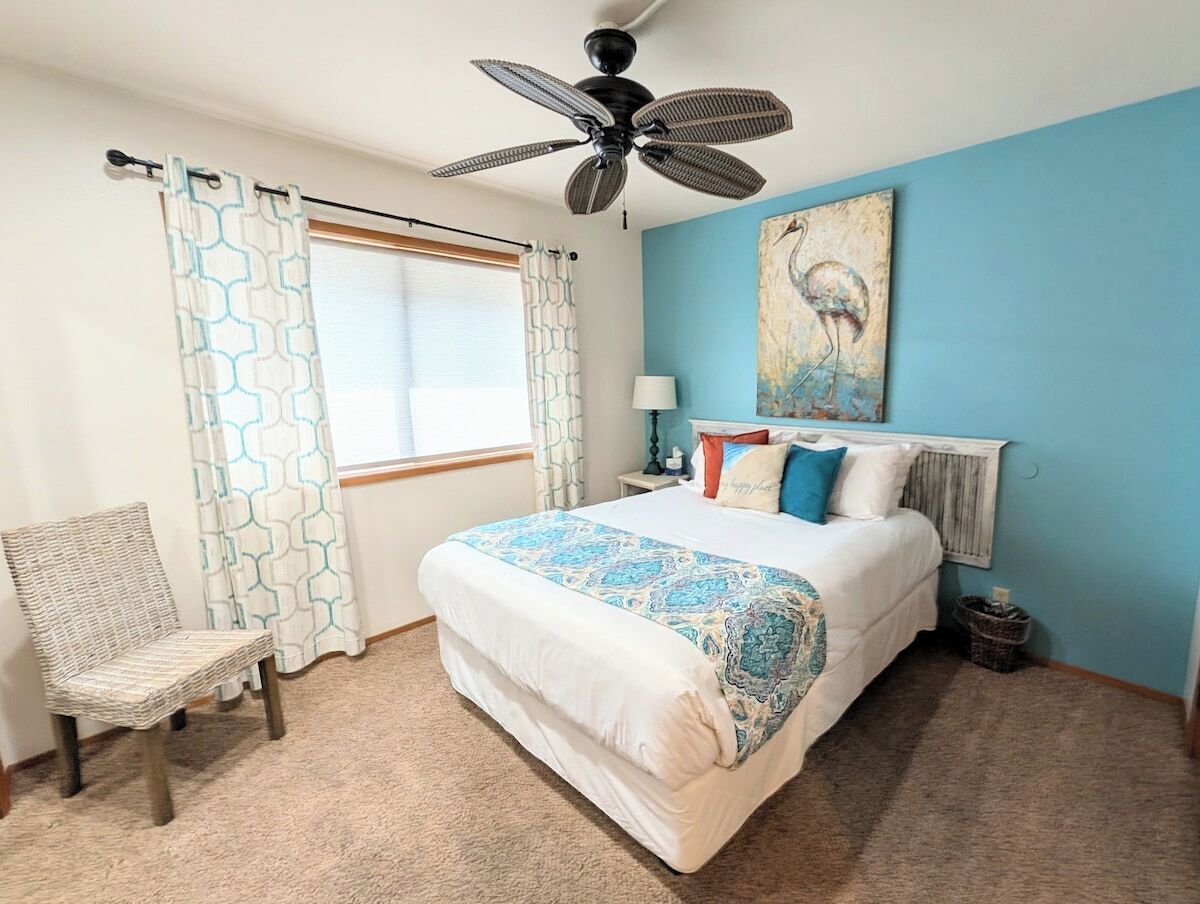 Un dormitorio con cama, una silla, ventilador de techo, ventana con cortinas y un cuadro en la pared.