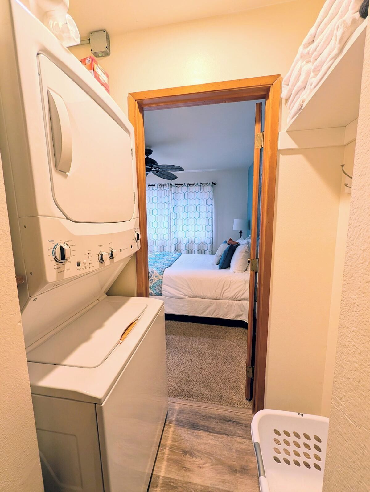 Un área de lavandería con lavadora y secadora, una cama con colcha de rayas azules y blancas, y una puerta de madera que da a un dormitorio.