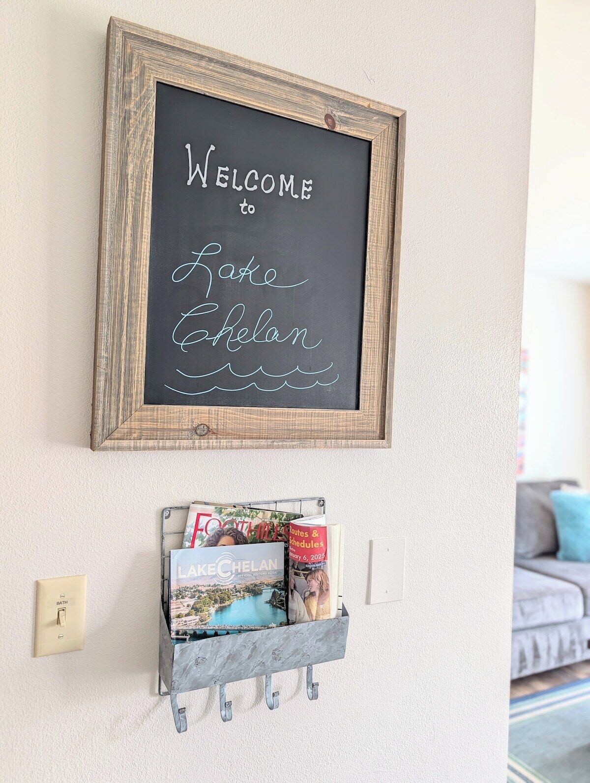 A welcome sign with a rustic wooden frame and chalkboard background, a small metal shelf below holding travel guides, and a light switch on the wall.