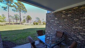 Une terrasse avec une table en verre et quatre chaises, un mur en briques et une vue sur un espace verdoyant avec des arbres et des bâtiments.