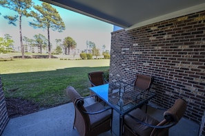 A patio with a glass table and four chairs, a brick wall, and a view of a grassy area with trees and buildings.