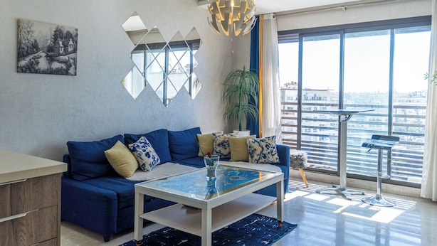 Un salon moderne avec un canapé bleu, une table basse en verre et une vue sur la ville grâce à de grandes fenêtres.