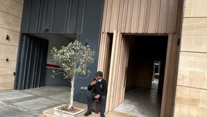 Un homme est assis devant un bâtiment, avec un pot de fleurs à proximité.