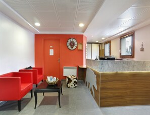 A reception area with a red wall, a clock, a reception desk, and a seating area with red chairs. - Cosy Apartment Near the Ski-slopes (Les Deux Alpes)