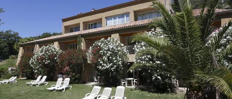 A two-story building with a balcony, surrounded by lush greenery and white flowering plants.