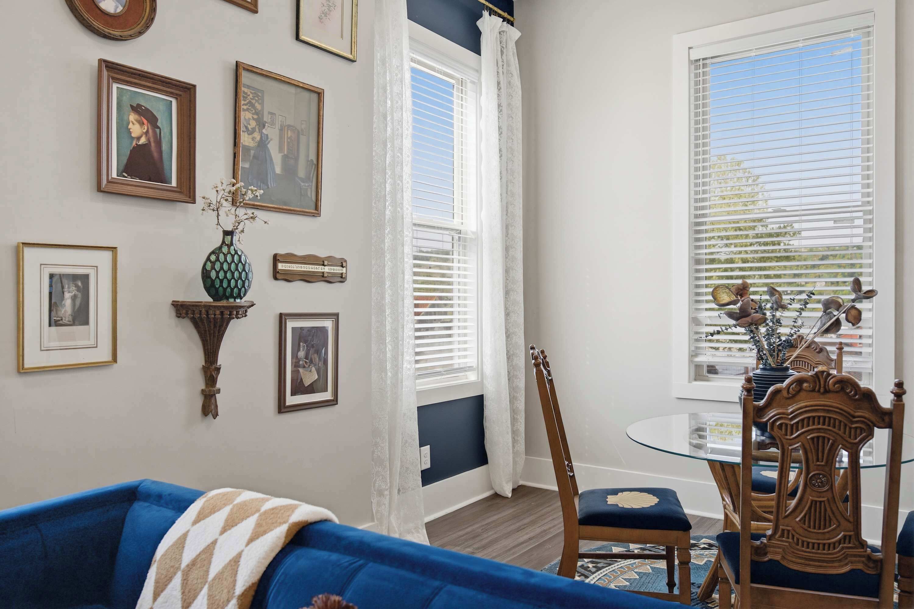 A living room with a blue sofa, a dining table with chairs, and a window with blinds.