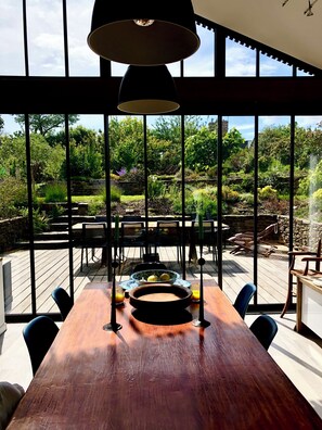 A dining area with a wooden table, chairs, and a view of a lush garden through large windows. - MiniTYCHA house-sea-garden-terrace-WIFI THD (Moëlan-sur-Mer)