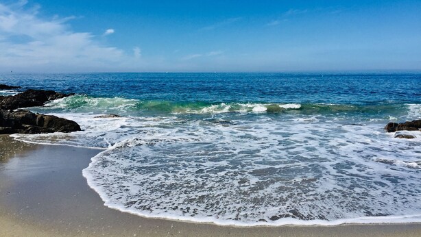 Ein Strand mit Wellen, blauem Himmel und felsiger Küste.