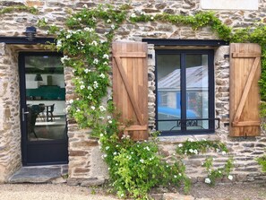 A stone building with wooden shutters, a glass door, and a window with a metal frame. - MiniTYCHA house-sea-garden-terrace-WIFI THD (Moëlan-sur-Mer)