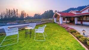 Un hotel con techo de tejas rojas, un césped con sillas y una mesa, y vistas a una carretera y árboles.