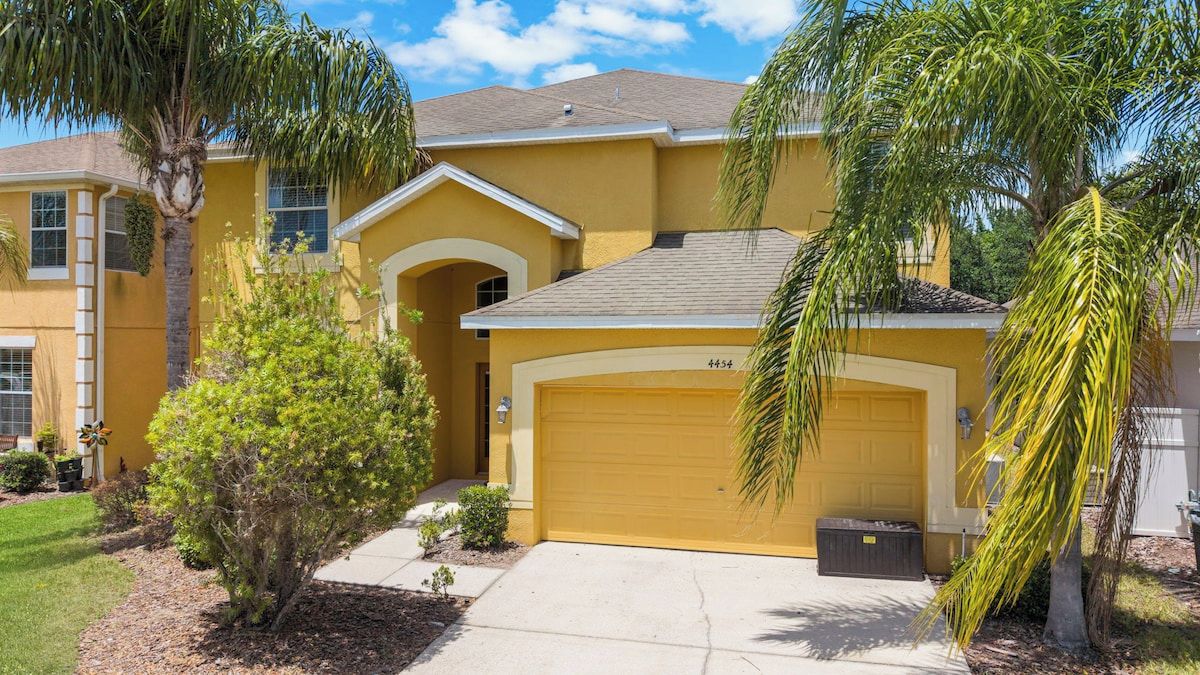 A yellow two-story house with a garage, surrounded by palm trees and a well-maintained lawn.