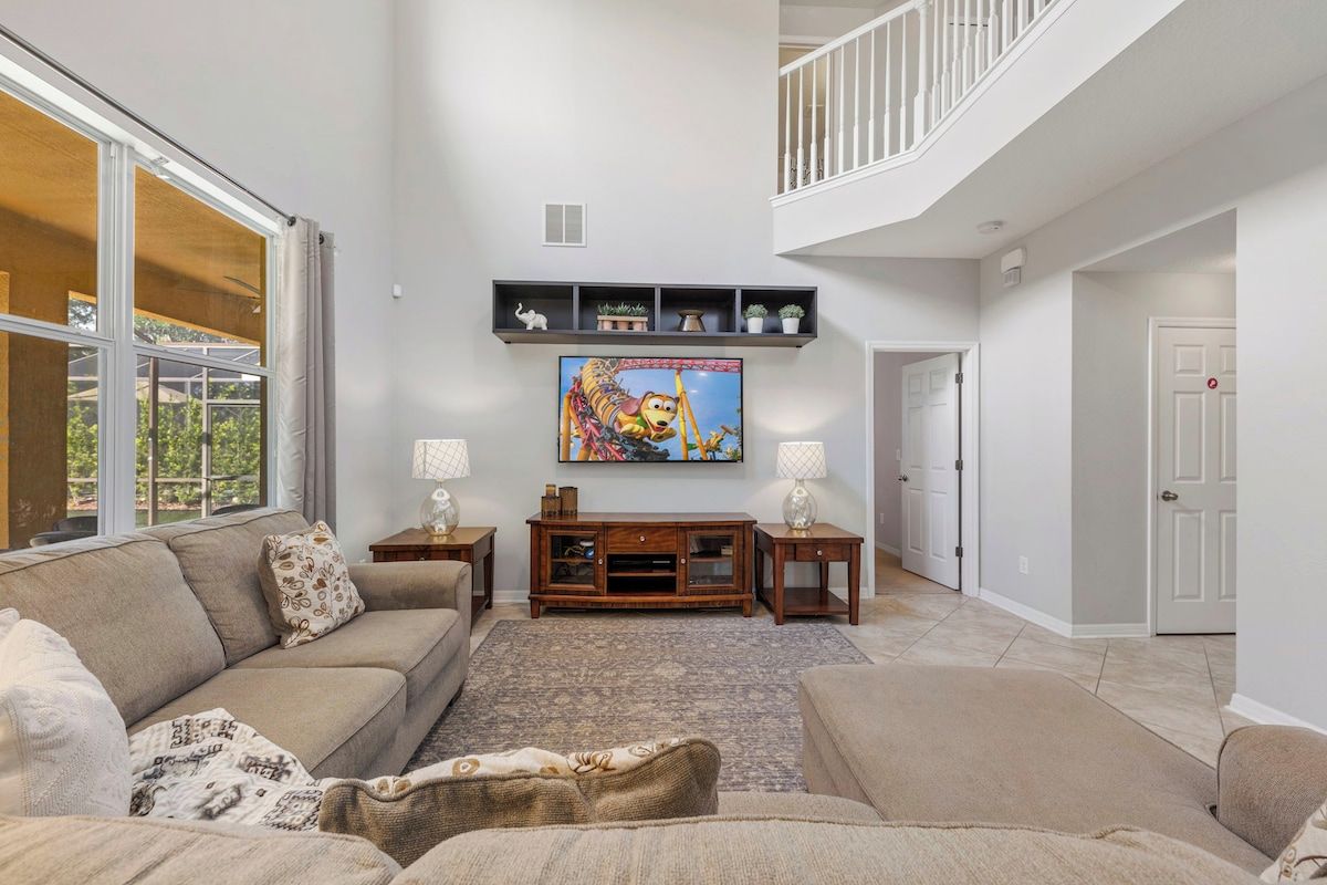A spacious living room with a high ceiling, a large window, a flat-screen TV, and a staircase leading to an upper level.