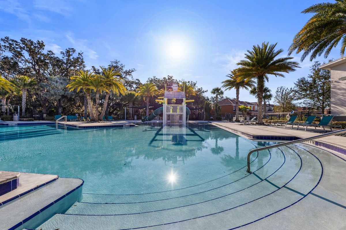 A swimming pool with a water slide, surrounded by palm trees and lounge chairs.