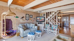 Ein gemütliches Wohnzimmer mit grauem Sofa, weißen Korbstühlen, einem Hängesessel und einem Fernseher, der 'Seaside Vacations & Sales' anzeigt.