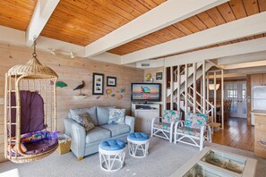 Ein gemütliches Wohnzimmer mit grauem Sofa, weißen Korbstühlen, einem Hängesessel und einem Fernseher, der 'Seaside Vacations & Sales' anzeigt.