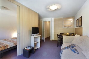 Quarto compacto com cama, televisão em móvel, sofá, escrivaninha e área de refeições.