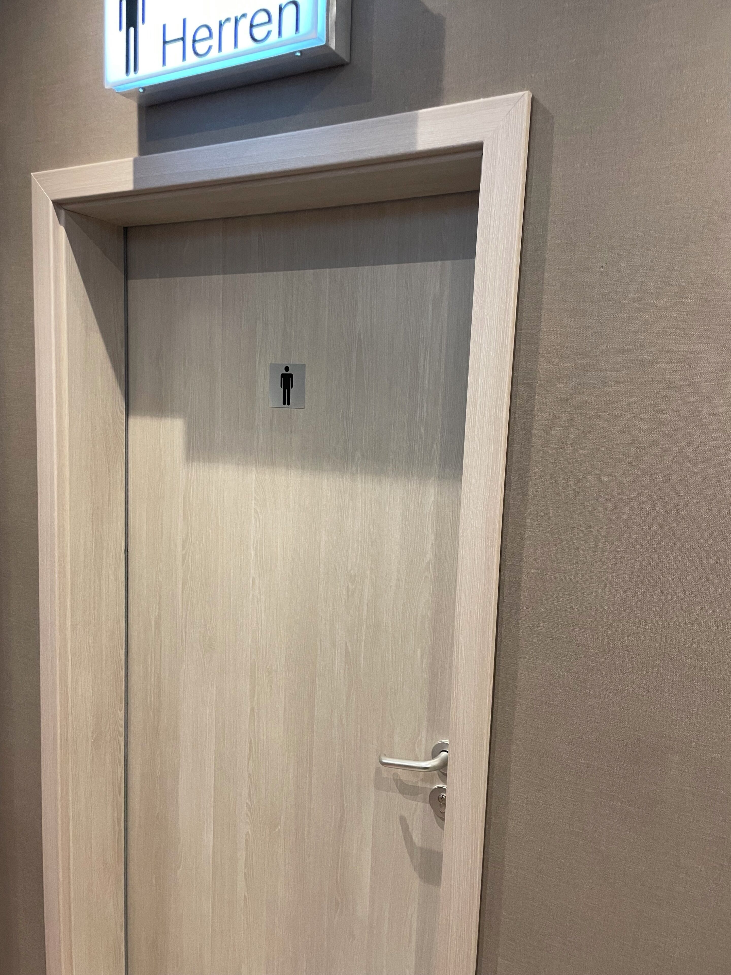 Uma porta de madeira com uma placa acima indicando 'Herren' e o símbolo de banheiro masculino na porta.
